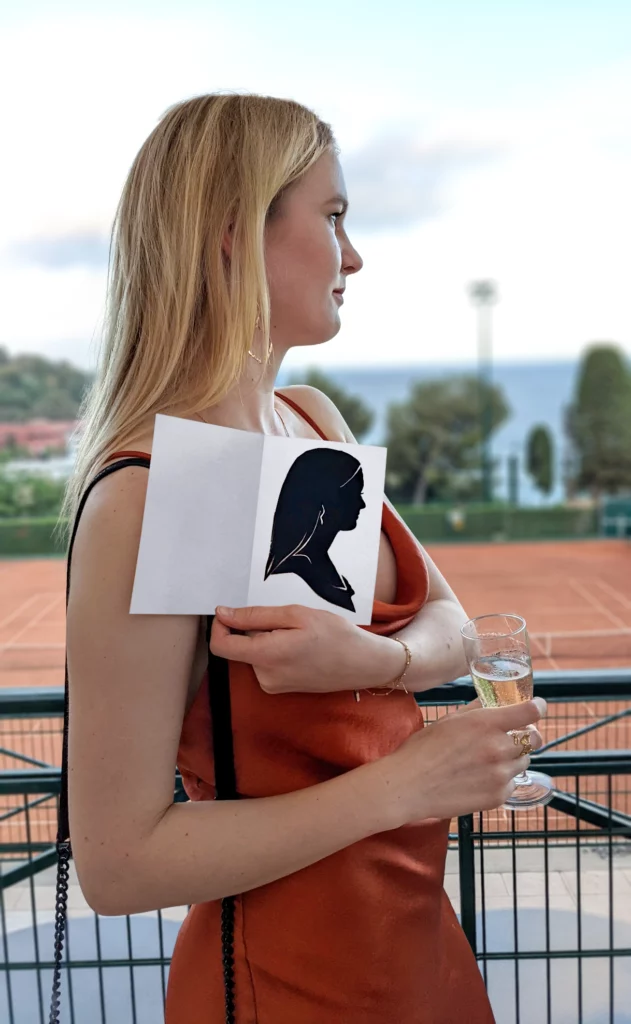Femme en robe rouge présentant son portrait découpé aux ciseaux par le silhouettiste Morlon devant les terrains de tennis de Roland Garros